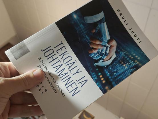 Kirja vinottain kädessä. Kansikuvassa kättelevät pukumies, jolla on robotin käsi, ja isosta näytöstä ulos kurottuva ihmisen käsi.