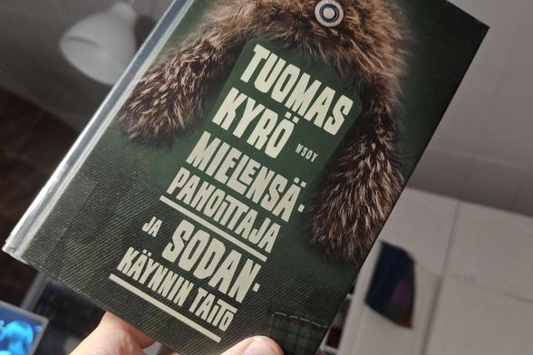Kirja kädessä. Kannessa kirjailijan nimen yllä Mielensäpahoittajan karvahattu, jossa sinivalkoinen sotilaan kokardi