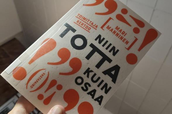 Kirja kädessä. Kannessa tekstien ympärillä oransseja välimerkkejä, pilkkuja ja huutomerkkejä
