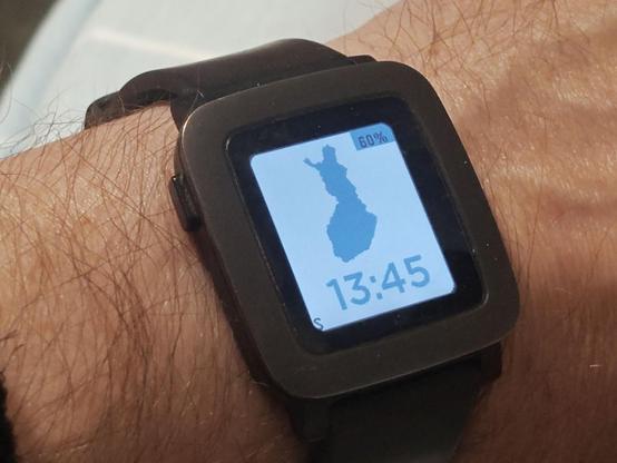 Ranteessa kello, jonka valkoisessa kellotaulussa on sininen Suomen muoto ja kellonaika.