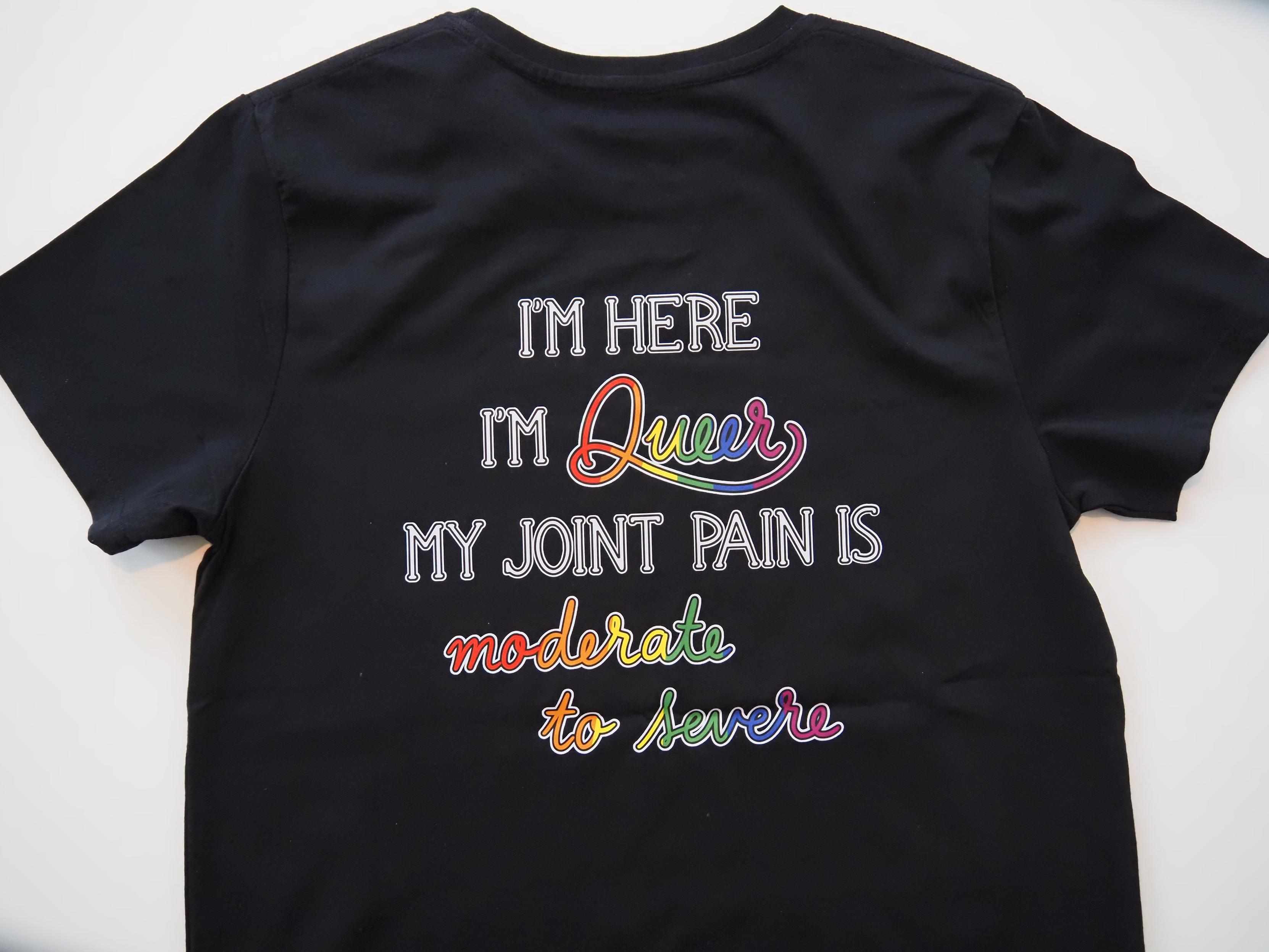 Kuva mustan t-paidan selkäpuolesta. Kuvana on teksti, jossa lukee I'm here, I'm Queer, my joint pain is moderate to severe. Teksti on muuten valkoista isoilla kirjaimilla mutta Queer ja moderate to severe on kirjoitettu kursiivilla ja väritetty sateenkaaren värein.