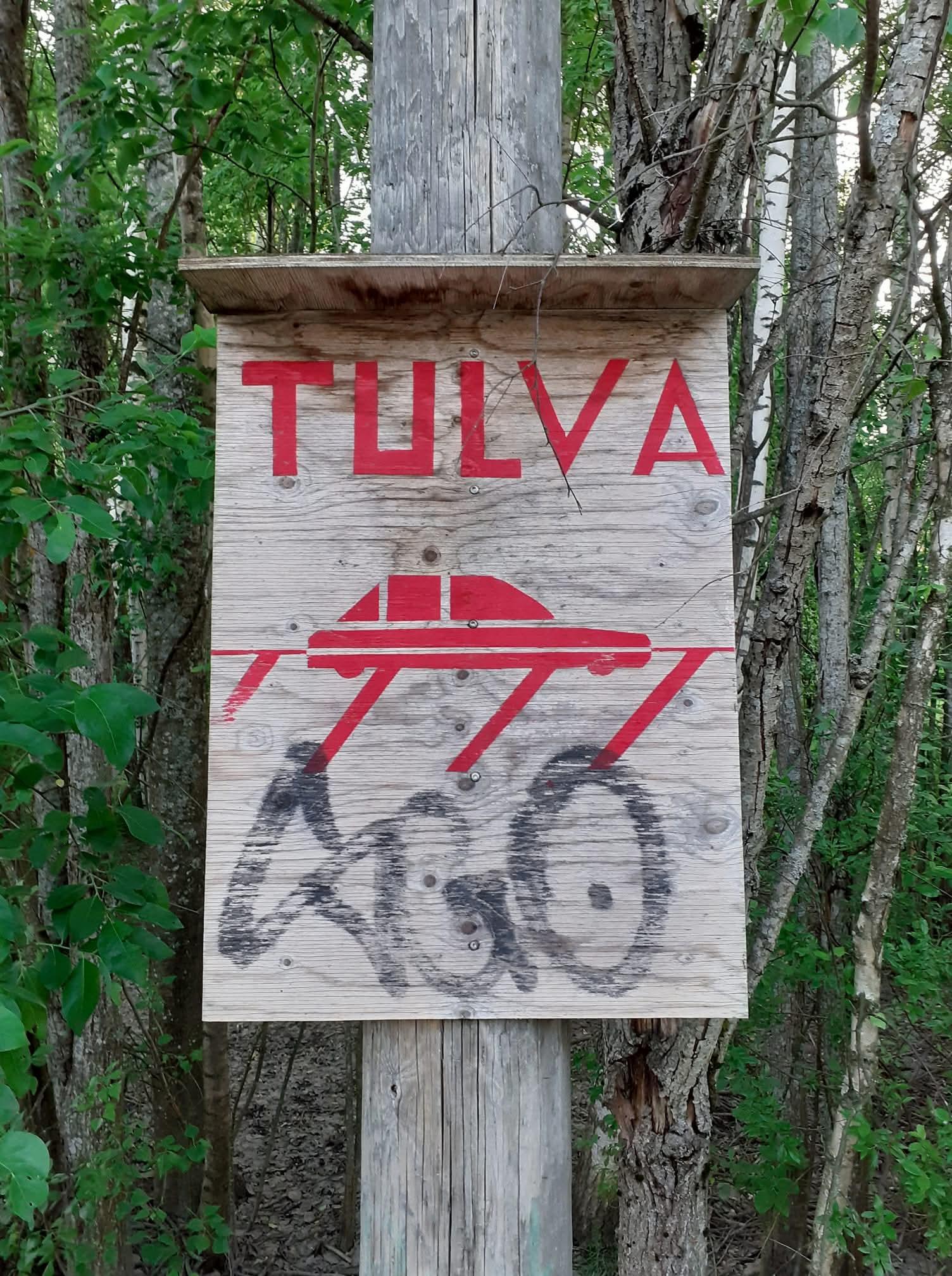 Vanerinen, ajan harmaannuttama kyltti, jossa on tyylitelty kuva autosta ja sen yläpuolella suurilla punaisilla kirjaimilla teksti TULVA. Tulvaa kuvataan vinoviivoilla, jotka ulottuvat auton konepellin tasalle. Tulvan alapuolelle kyltin alaosaan on joku piirustanut täginsä.
