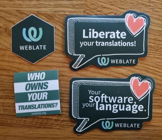 A collection of Weblate stickers on a wooden desk. Hexagonal with Weblate logo, two with comic-bubble styled stating “Liberate your translations!” and “Your software, your language!”. The last is Nextcloud-themed with a motto “Who owns your translations?”