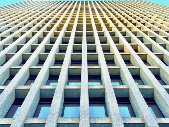 A view from the sidewalk up at the Standard Insurance Center in Downtown Portland reveals vast concrete framing and seemingly endless vertical windows beneath a clear blue sky.