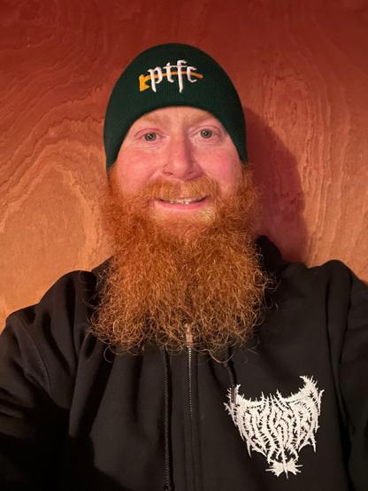 Man with a gorgeous ginger beard wearing a black hoodie with the Timbers metal logo on the chest and a green beanie with the PTFC Gothic Axe logo, grinning at the camera.