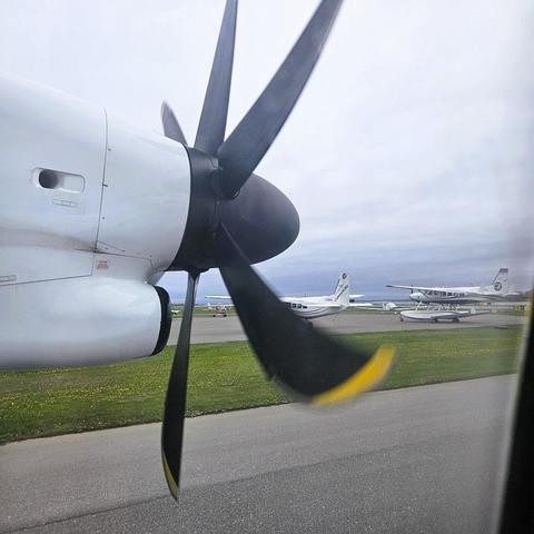 View out the window of an airplane at the propeller and some small planes including a float plane on wheels on the tarmac