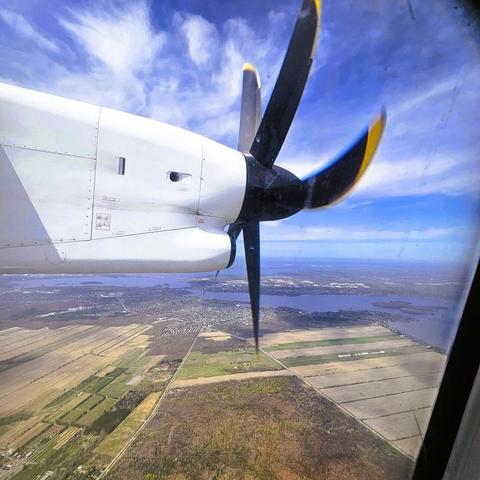 View out the window of a plane with the propeller in view, looking down on a patchwork of fields and a river.