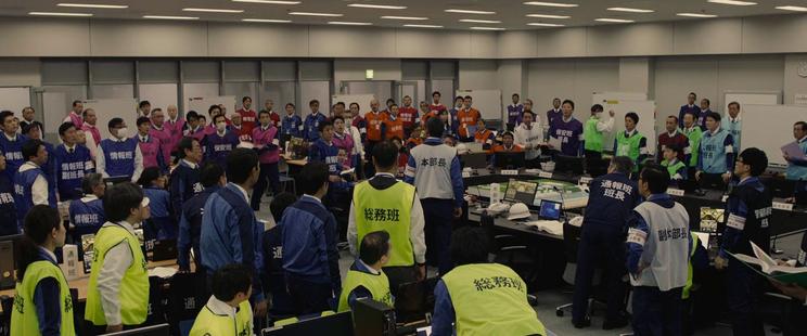 A large room full of people looking at a person in the center making an announcement. Everyone is wearing colour-coded vests that describe their role in large Japanese letters.