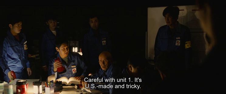 Several people in blue overalls sitting around a table in a dark room lit with an emergency lamp on a table. There's a large manual open on the table. An old, bald man is speaking. The subtitle reads "Careful with Unit 1. It's U.S.-made and tricky."
