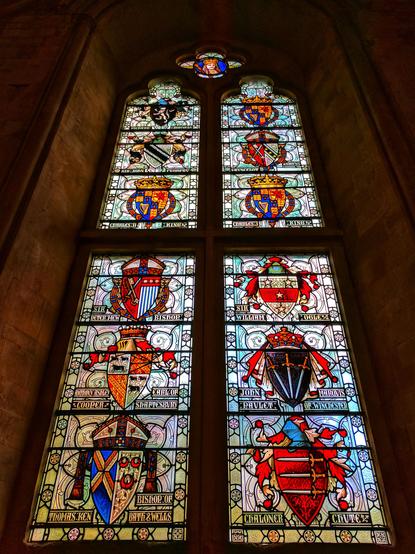 A stained glass window in the Great Hall, Winchester. The window is tall and narrow, with multiple panes. Each pane contains a different heraldic shield, with colourful designs and inscriptions. The shields are arranged in a grid pattern. The background is a dark, stone wall.