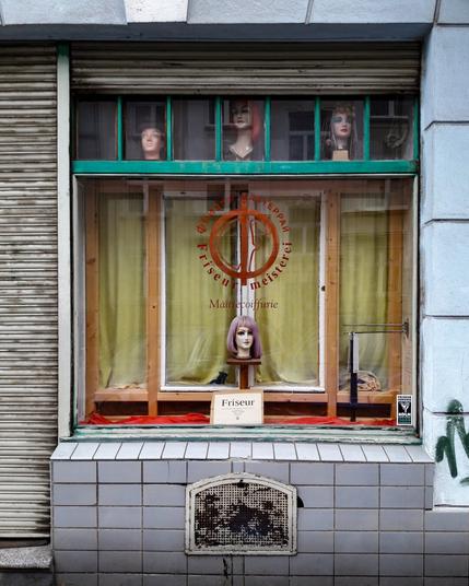 Friseursalon Köln: Oben drei Puppenköpfe (dunkel gelockt, rot hochgesteckt, blond gewellt mit blauem Band) auf grünem Sims. Unten mittig lila Kurzhaar-Puppe vor gelben Vorhängen. Orangefarbenes Logo "Friseurmeisterei/Maître coiffure" auf der Scheibe. Weißes Schild "Friseur" davor. Roter Stoff am unteren Rand. Grauer Fensterrahmen, darüber graues Rolltor. Graue Fliesen unter dem Fenster, darunter dunkler Gitterrost. Rechts graue Wand mit Graffiti und kleinem Vogel-Aufkleber.