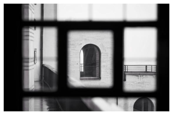 Schwarz-Weiß-Foto im Querformat mit weißem Rand. Durch ein in kleine Felder unterteiltes Fenster, dessen Rahmen nur unscharf zu sehen sind, blickt man auf ein scharfes, weiter entferntes, rundbogenförmiges Fenster, durch das man wiederum hindurchsehen kann. Das zweiter Fenster ist so genau in den Rahmen des ersten hineinfotografiert, dass es einen Rahmen ergibt.