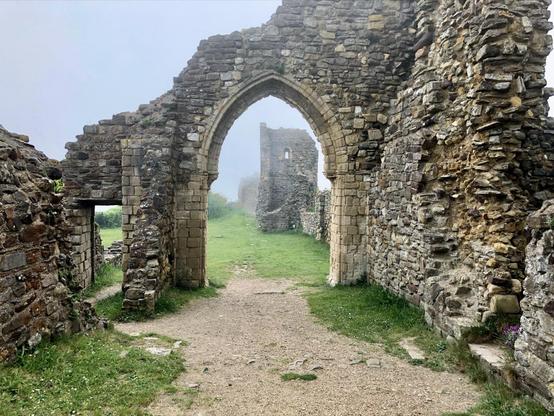 The image shows the remains of an old stone structure, likely a ruins of a medieval building or castle. It features an arched gateway through which a grassy path leads. The archway is made from various sizes of stone and part of its surrounding wall is also visible, which are in various states of decay. The environment appears foggy or misty, adding an atmospheric and somewhat mysterious ambiance to the setting. In the distance, another part of the ruins can be seen, suggesting the site might have once been a larger complex. The ground is covered in grass and the path appears well-trodden, indicating it might be a place visited by tourists or hikers.