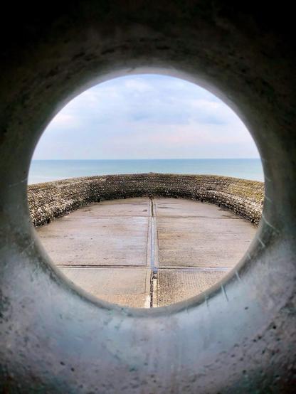 This image shows a view through a circular opening, possibly a pipe or a tunnel, opening out to a scenic view of the ocean. The foreground has a textured, rounded surface, perhaps a wall or barrier made of concrete or stone. Directly in front, the landscape includes what looks like a walkway or a ramp leading toward the ocean. The design of the barriers along the pathway is accentuated with pebbles or cobblestones. The horizon where the sky meets the sea is visible in the distance, creating a calm and serene scene. The framing through the opening adds an artistic touch, focusing the viewer's eye directly towards the center of the image.