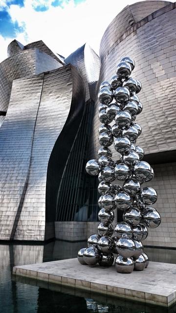 A tall sculpture made of numerous shiny, reflective metal spheres stands on a stone platform in front of the textured titanium walls of the Guggenheim Museum Bilbao. The sculpture reflects the sky and surrounding architecture.