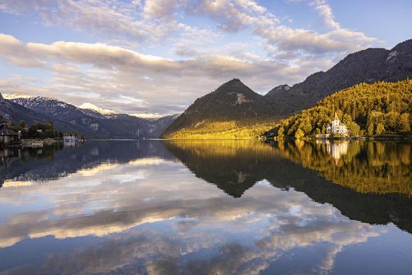 Symmetrical reflection shot of Grundlsee lake in Austria as sunset, under a blue sky with pinkish cloud bands while the sun is painting a yellow glow on lower parts of the mountain.