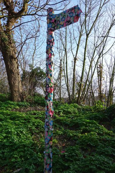 A signpost, covered in colourful patterned fabric and small round stickers, stands in a patch of green ground cover. The signpost has a single directional arrow pointing to the right. Behind it are bare trees against a blue sky. A wire fence is visible in the mid-ground. The fabric on the post and arrow features a repeating floral and geometric pattern with pink, blue, and white as prominent colours. The round stickers are scattered across the signpost.