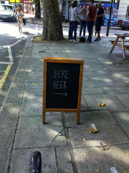A black A-frame pavement sign with a wooden frame displays the words "BERE HEER" in white lettering, with a right-pointing arrow underneath. The sign stands on a grey pavement, with some fallen leaves scattered around it. In the background, a group of people are standing and talking on the pavement next to a tree, and a cyclist is riding past. A street and buildings are visible further in the background.