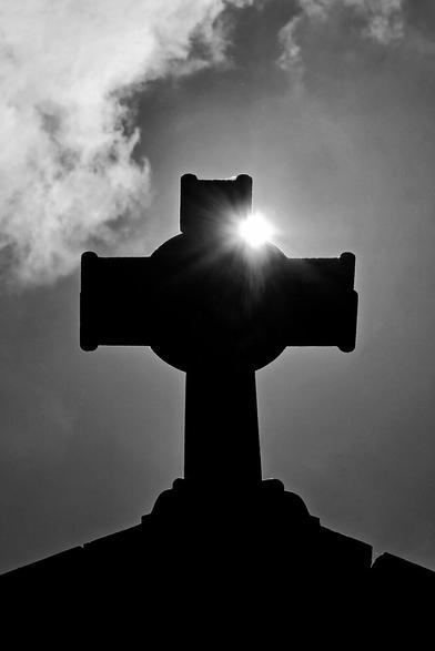 A black and white, low-angle shot shows a silhouetted Celtic cross against a bright sky where the sun is shining from behind the center of the cross.