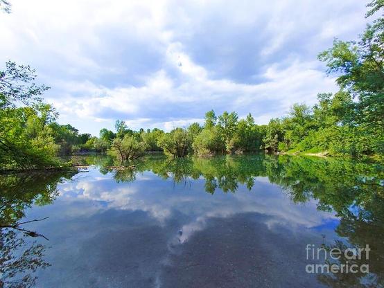 A serene lake reflects the lush greenery and cloudy sky overhead, creating a symmetrical and peaceful scene. Trees line the far shore, and their vibrant green hues contrast beautifully with the deep blue water.
Reflections on the Lake in Bundek Park, Zagreb