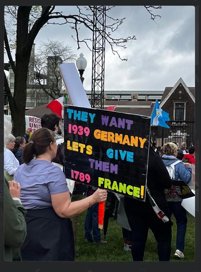 The image shows a demonstration with a protester holding a banner with the words -

“ THEY WANT
1939 GERMANY
LETS GIVE
THEM
1789
FRANCE!”