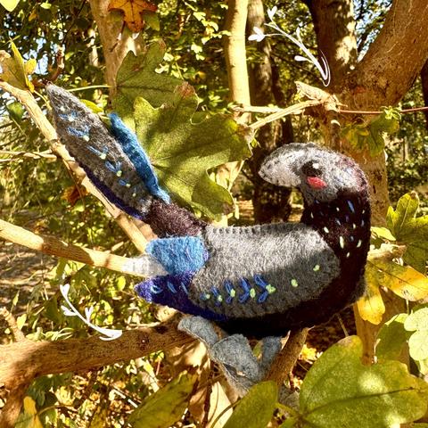 In the branches of a sunlit tree is standing a groove-billed ani felt plushie. It has a grey head and beak and a black body. It’s wing is grey with two-toned blue feathers coming out of, it’s tail looks similar.  On its neck, wings and tail, blue and green embroidery curve along the body.