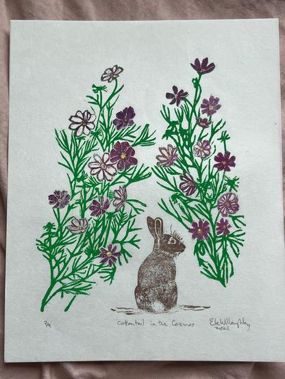An ink print on a white paper. In the forground is a realistic looking brown rabbit. It looks like it is about to enter a tangle of purple and white flowers with green stems. The print is titled "Cottontail in the Cosmos" and it is print 7/9 and was made by Ele Willoughby in 2021.