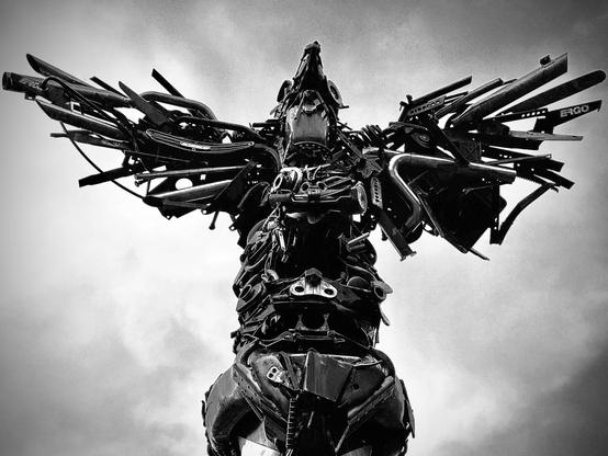 Artist Travis Pond's "Wolf, Beaver, Orca, Frog, Raven" totem pole sculpture stands tall against a cloudy sky, crafted from repurposed automotive steel parts to form different notable animals.