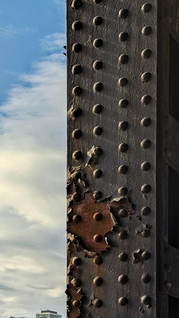 A partly cloudy day with a steel post, covered in dozens of rivets along its face, painted over in black, and a section towards the lower 1/3rd peeling off, exposing oxidized (rusted) iron.