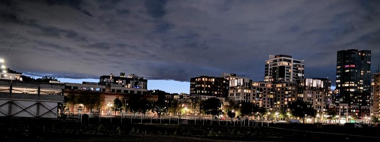 A panoramic shot overlooking a multi-acre empty lot, with a dozen high rise condos and apartments lit up, with an angry, cloudy sky, horizontally demarcated off in the horizon by a clear sky.