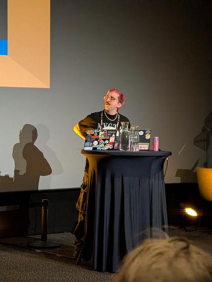 @errs on stage looking at the screen. Sporting short purple hair, a moustache and glasses. Wearing a black t shirt.