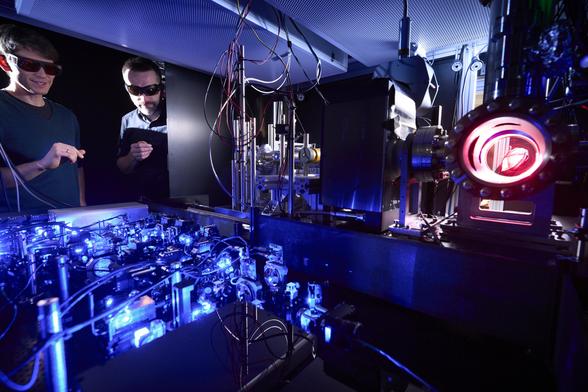 Two people - Johannes Zeiher and Sebastian Blatt - monitor an experiment at the Max Planck Institute of Quantum Optics. You can see a lot of lasers and blue light. The scientists are wearing glasses. Credit: © Max Planck Institute of Quantum Optics / Munich Quantum Valley
