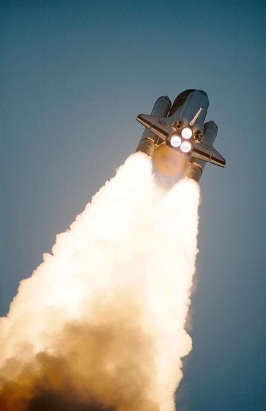 A space shuttle takes off into a gray-blue sky, leaving a bright column of white vapor behind it. The view is from below.