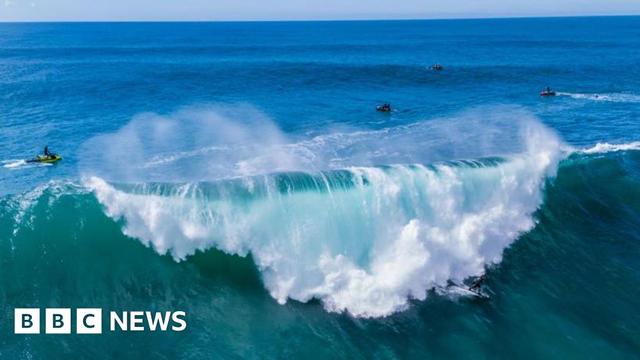 A large wave in the Atlantic breaks. The water is a deep blue green colour. There is a surfer and people on jetskis.