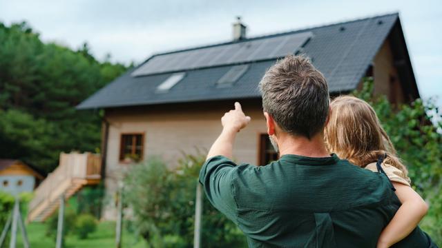 Un père de dos tient sa petite fille dans ses bras et lui pointe les panneaux solaires sur le toit d'une maison.