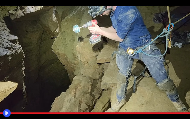 Speleologo si sporge sopra un baratro oscuro in un ambiente sotterraneo, tenendo in mano un trapano con un rocchetto di filo all'estremità.