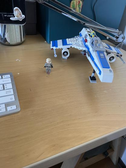 Su una scrivania di legno chiara, c'è un piccolo modellino Lego di un personaggio femminile, con capelli biondi e un'uniforme beige. Accanto a lei, c'è un caccia stellare Lego bianco e blu, con dettagli intricati.  Un robottino di Lego giallo è visibile nella cabina di pilotaggio del caccia.