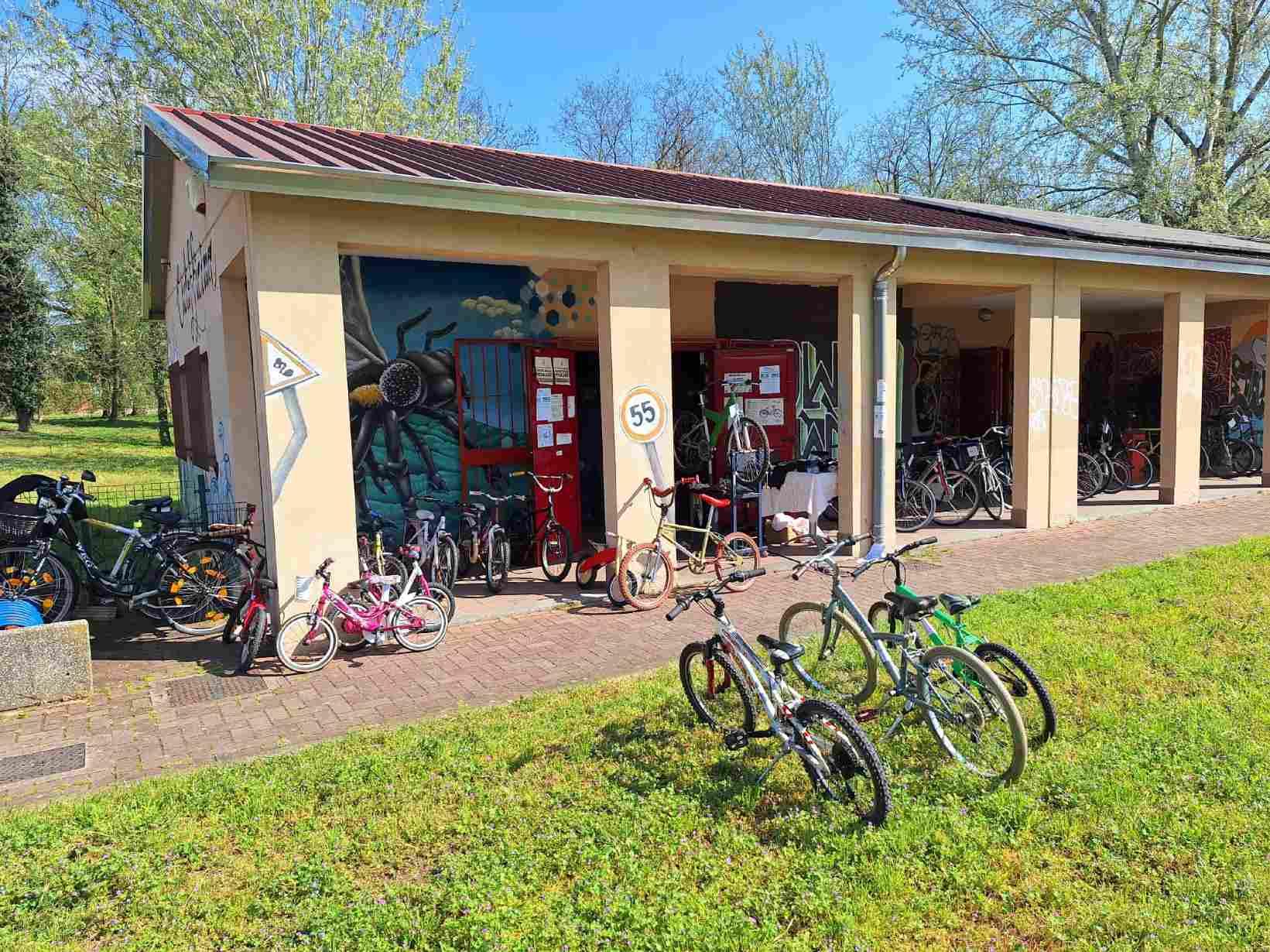 Foto della ciclofficina vista dal prato di fronte. Si tratra di un edificio ad un piano con un porticato. Ci sono alcune bici dell'asta sul prato