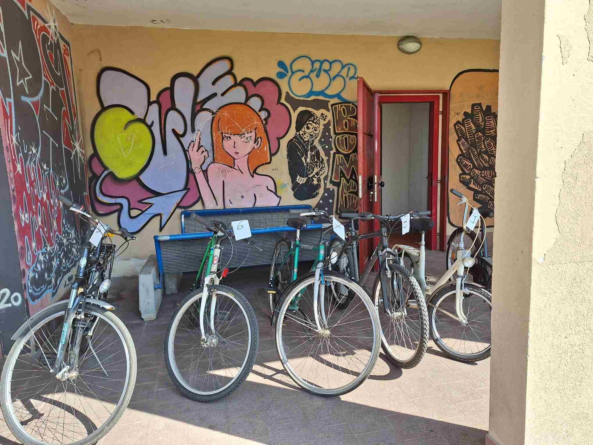Foto di 5 bici sotto al porticato della ciclofficina pronte per essere battute. Sui muri ci sono alcuni murales