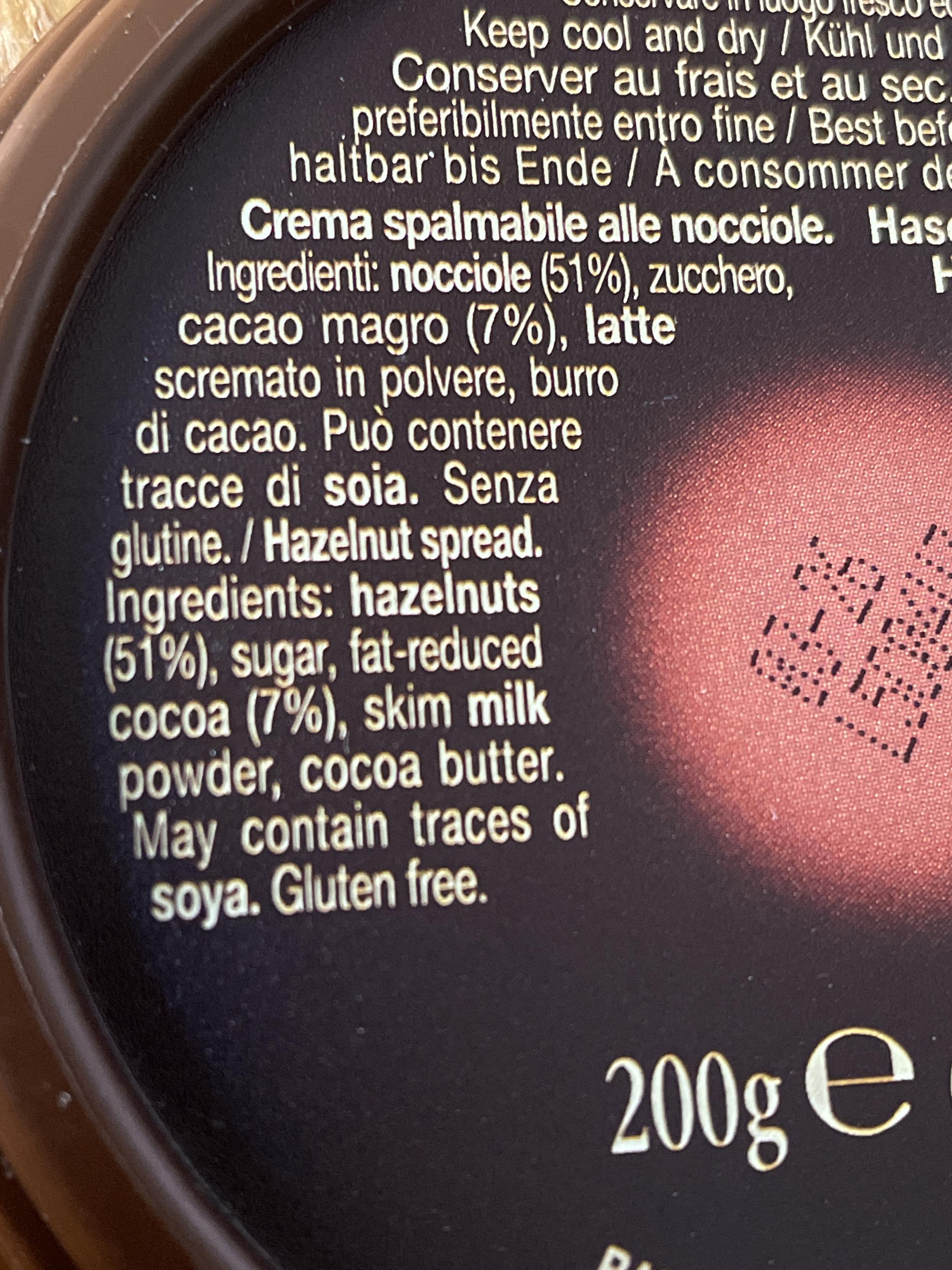 L'immagine mostra l'etichetta di un prodotto alimentare. L'etichetta ha un retro bianco con testo scritto in diverse lingue. In alto c'è scritto: "Conserver cool et dry / Kühl und trocken / Keep cool and dry". Subito sotto, in caratteri più piccoli: "À consommer de préférence avant le / Bis zum / Best before". La parte inferiore dell'etichetta include una lista di ingredienti: "Nocciole (51%), latte scremato (7%), zucchero, burro". Il testo riporta anche: "Può contenere tracce di soia / Senza glutine". Alla fine, c'è scritto "200g".