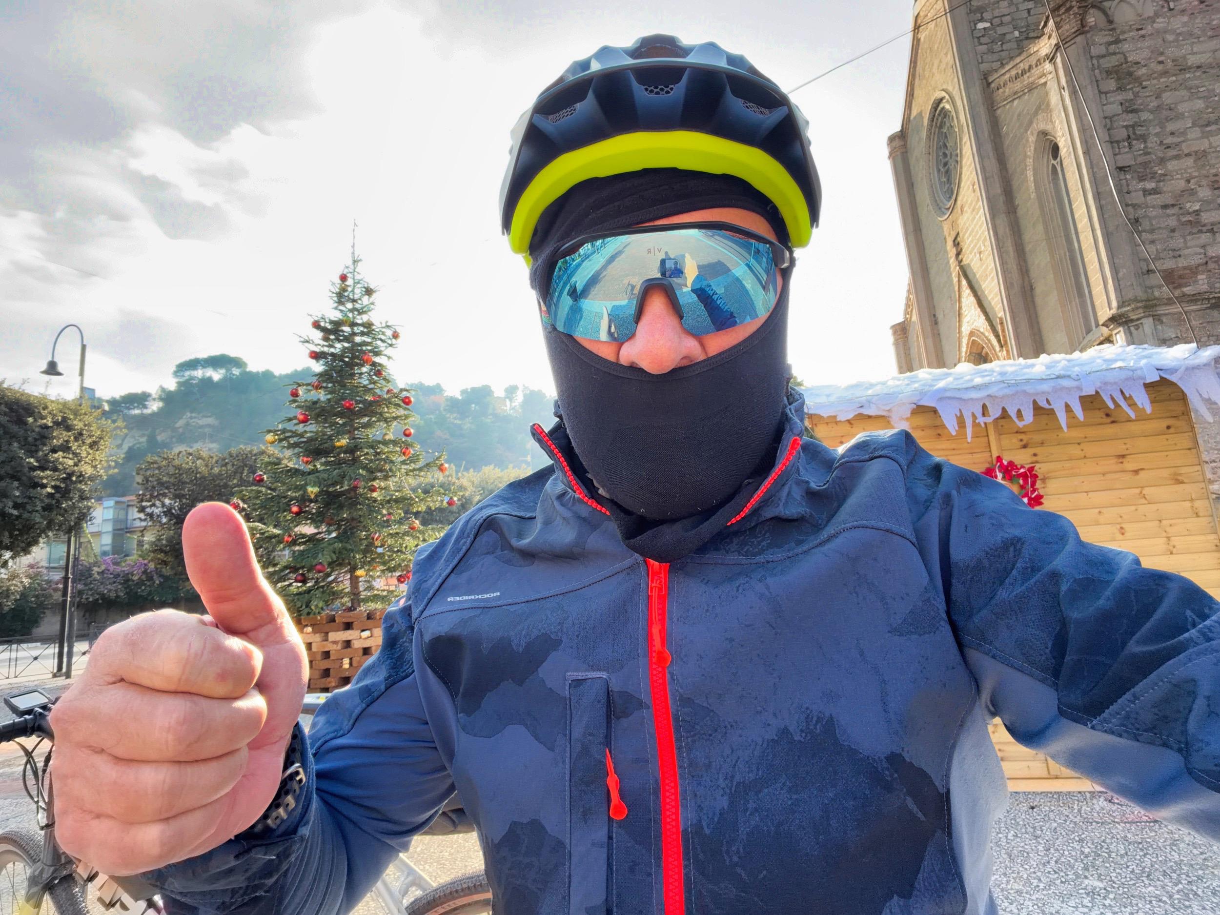 A cyclist wearing a black face mask, helmet, and sunglasses gives a thumbs-up. In the background, there is a decorated Christmas tree and a building with architectural details. The scene is outdoors on a sunny day