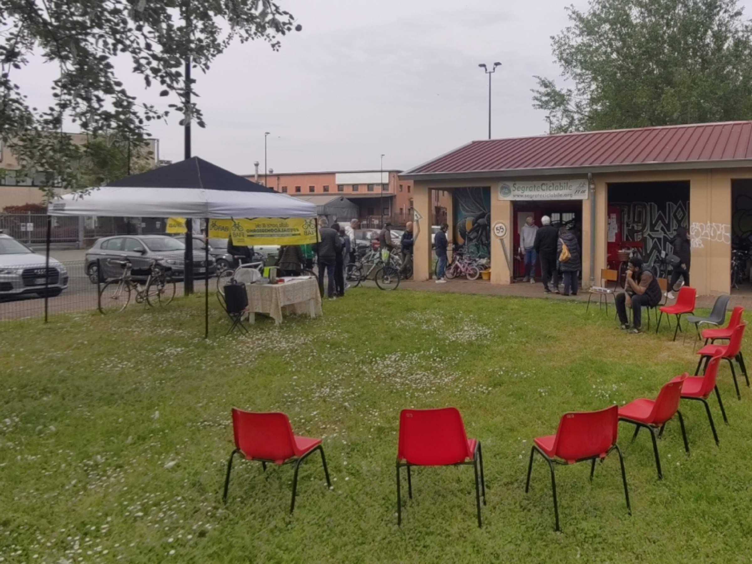 Foto del presto esterno della ciclofficina. Un gazebo di SegrateCiclabile co varie sedie intorno. Sotto al porticato numerose bici e persone che guardano e le provano