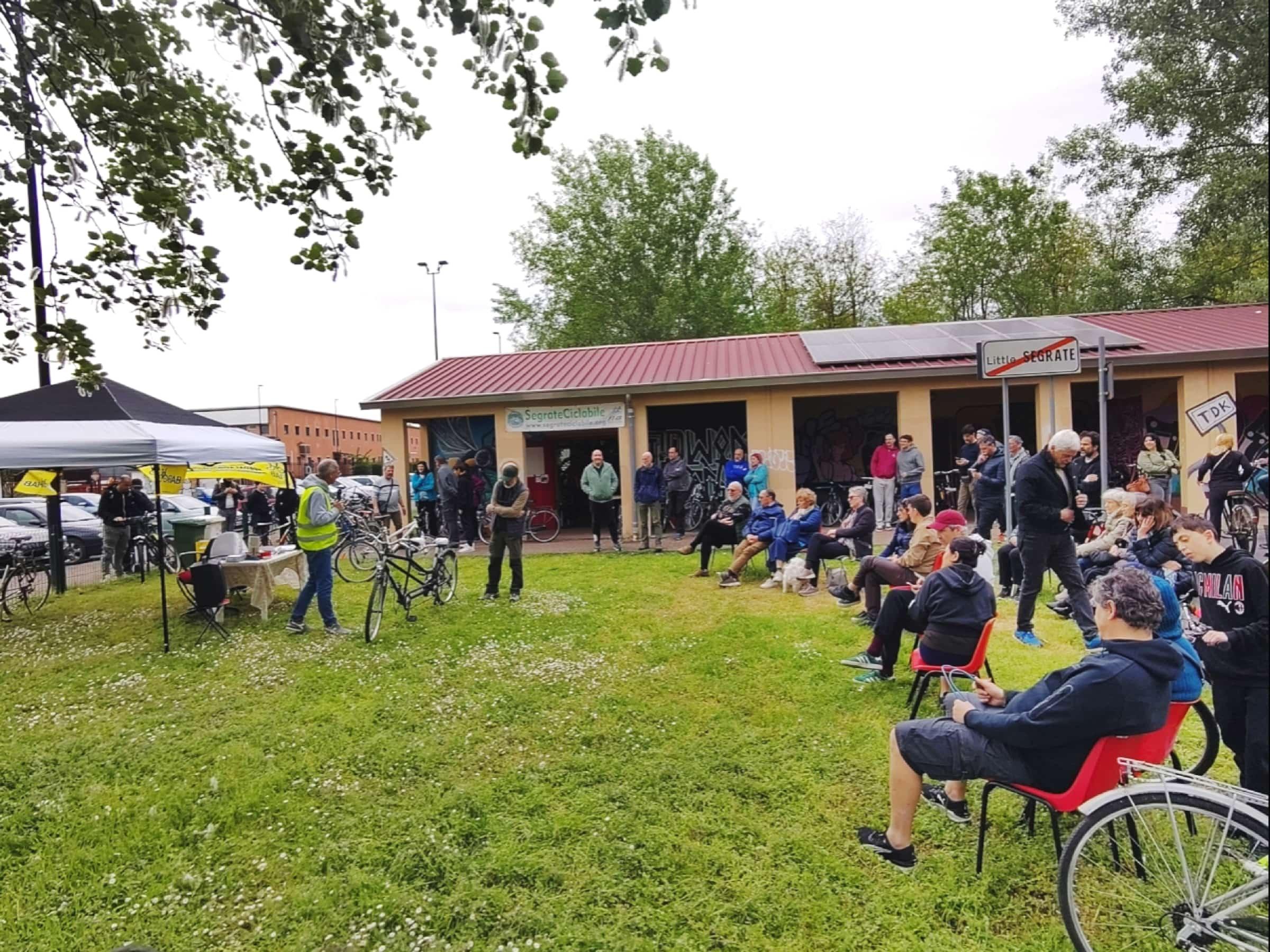 Foto del prato esterno della ciclofficina. Un gazebo di SegrateCiclabile con varie persone sedute intorno. Un socio attivo con un microfono che presenta una bici durante l'asta.