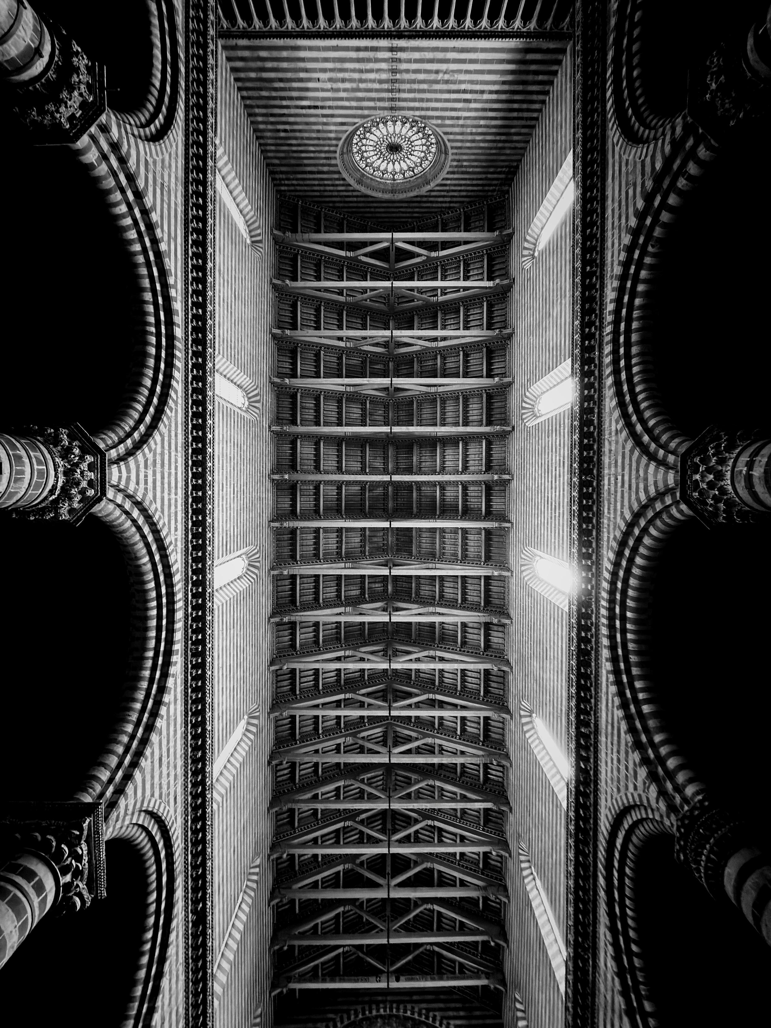 La fotografia mostra il soffitto del Duomo di Orvieto, ripreso dal basso verso l’alto in bianco e nero. L’inquadratura mette in evidenza l’architettura gotica, con archi a tutto sesto e colonne laterali ornate. Le travi e le nervature del tetto creano una sequenza ritmica e geometrica, mentre al centro si nota un rosone circolare. La luce filtra lateralmente, accentuando i contrasti tra le zone illuminate e le ombre profonde.
