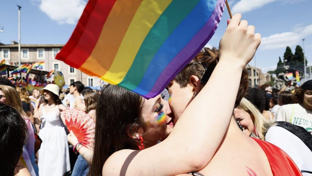 Nella fotografia, due ragazze abbracciate mentre si baciano. Una delle due ha in mano una bandierina col colori dell'arcobaleno.