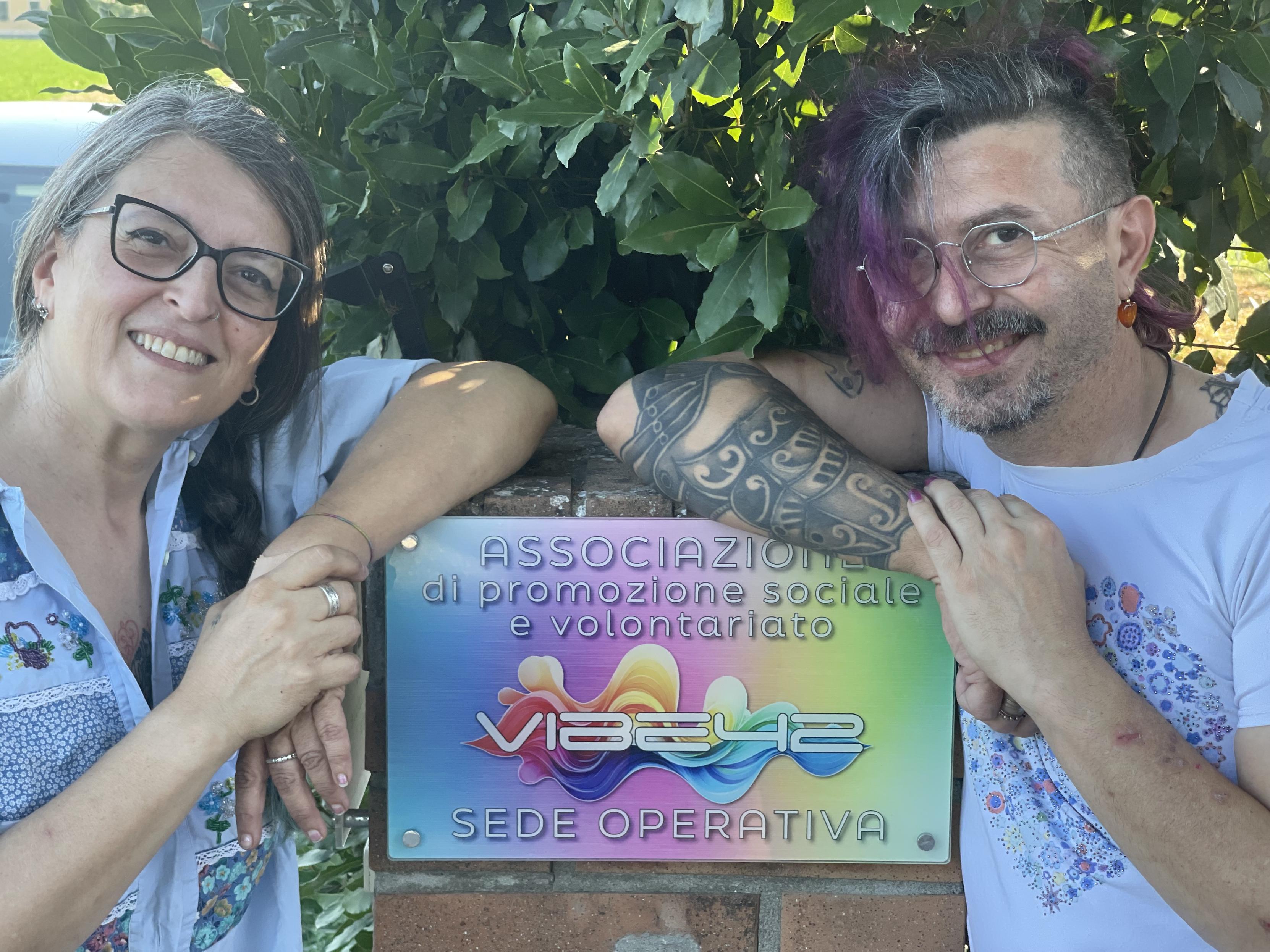 Due persone sorridenti sono riprese in primo piano all’aperto, ai lati di una targa dell’associazione Vibe42 fissata su un muretto di mattoni. Dietro di loro c’è una folta siepe verde; la scena trasmette un’impressione informale, accogliente e concreta.

A sinistra c’è una donna con occhiali neri e capelli grigi raccolti, che sorride appoggiando il braccio sulla targa. A destra c’è un uomo con capelli scuri con ciocche viola, occhiali sottili, barba e un tatuaggio visibile sul braccio; anche lui sorride e si appoggia alla targa. Al centro, la targa colorata riporta il nome Vibe42 e la dicitura “associazione di promozione sociale e volontariato”.