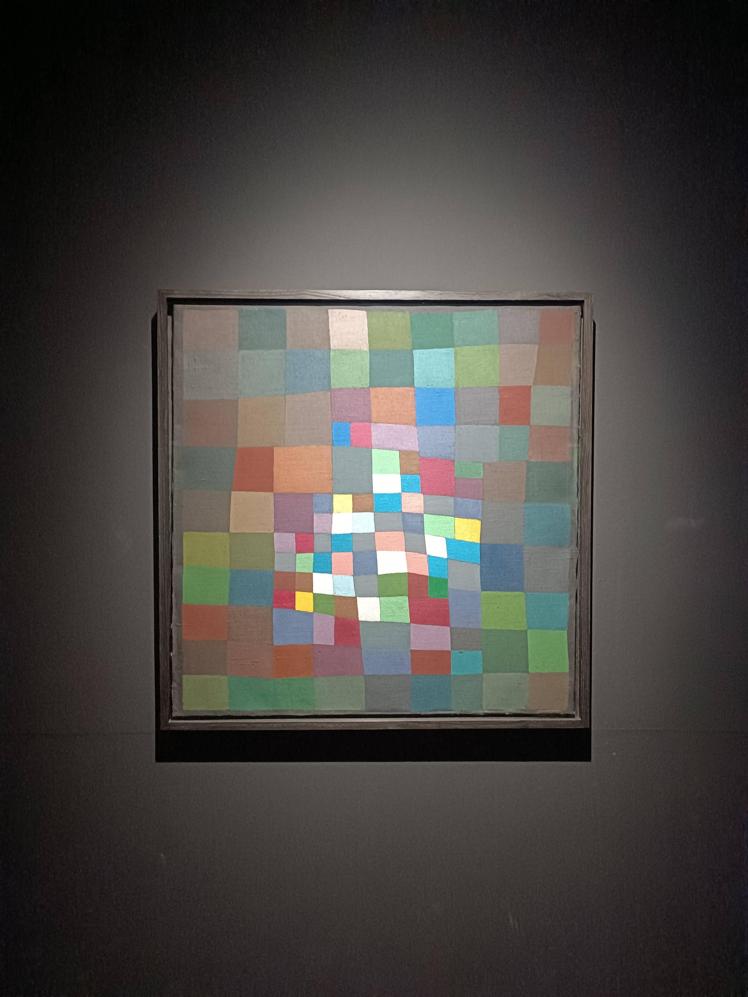 Un dipinto astratto quadrato con una cornice scura è appeso su una parete scura. Il titolo è "Flowering", dipinto da Paul Klee. L'opera consiste in numerosi piccoli quadrati colorati disposti in un motivo simile a una griglia. Al centro della composizione, si trova un gruppo di quadrati bianchi, azzurri e rosa chiaro. Verso i bordi, le tonalità passano a varie sfumature di verde, blu, rosso e marrone. L'opera è illuminata da una luce proveniente dall'alto che mette in risalto il quadro rispetto allo sfondo scuro.