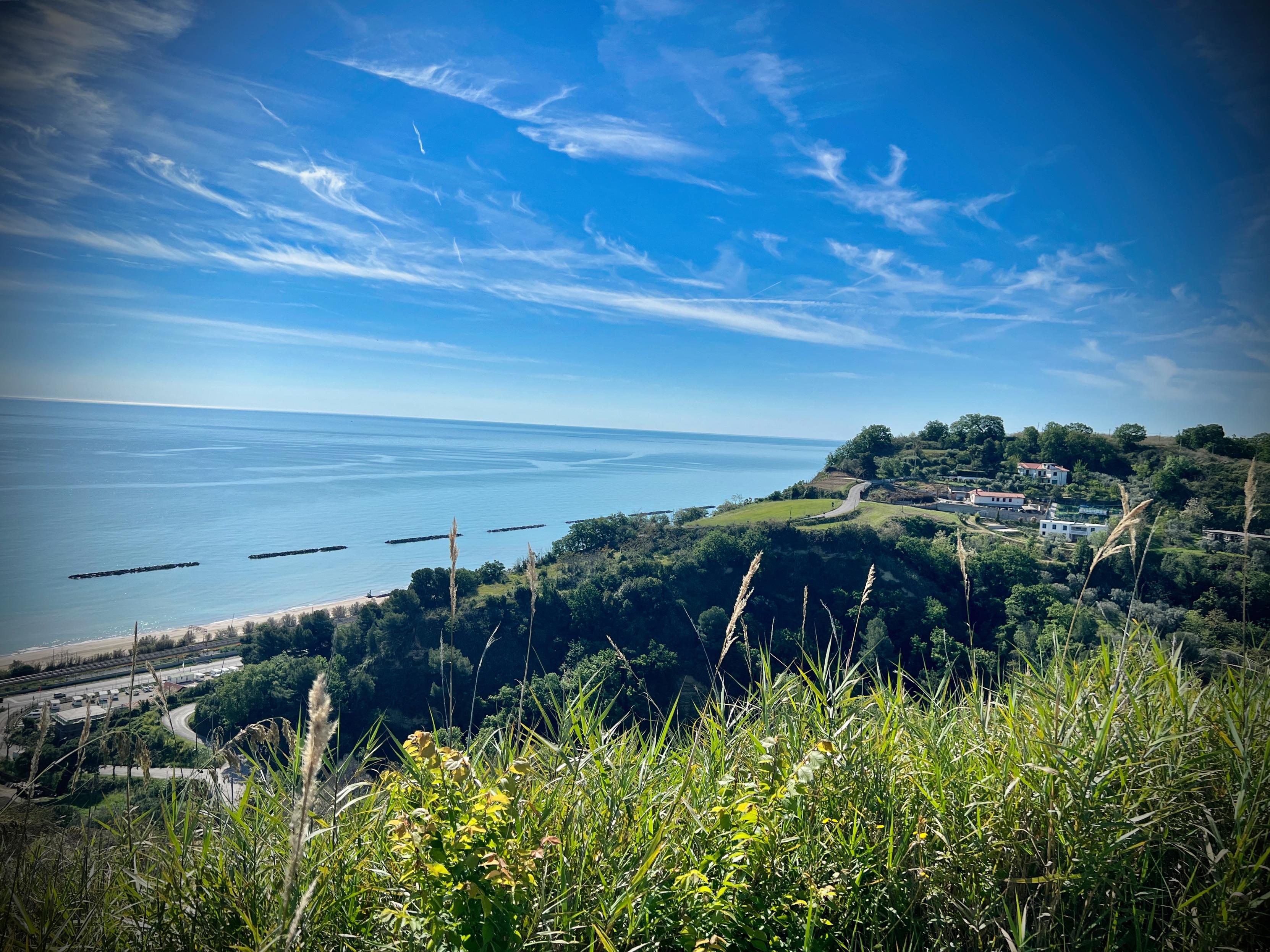 Paesaggio costiero con mare blu, cielo azzurro e nuvole bianche, vista di colline verdi con case e vegetazione.