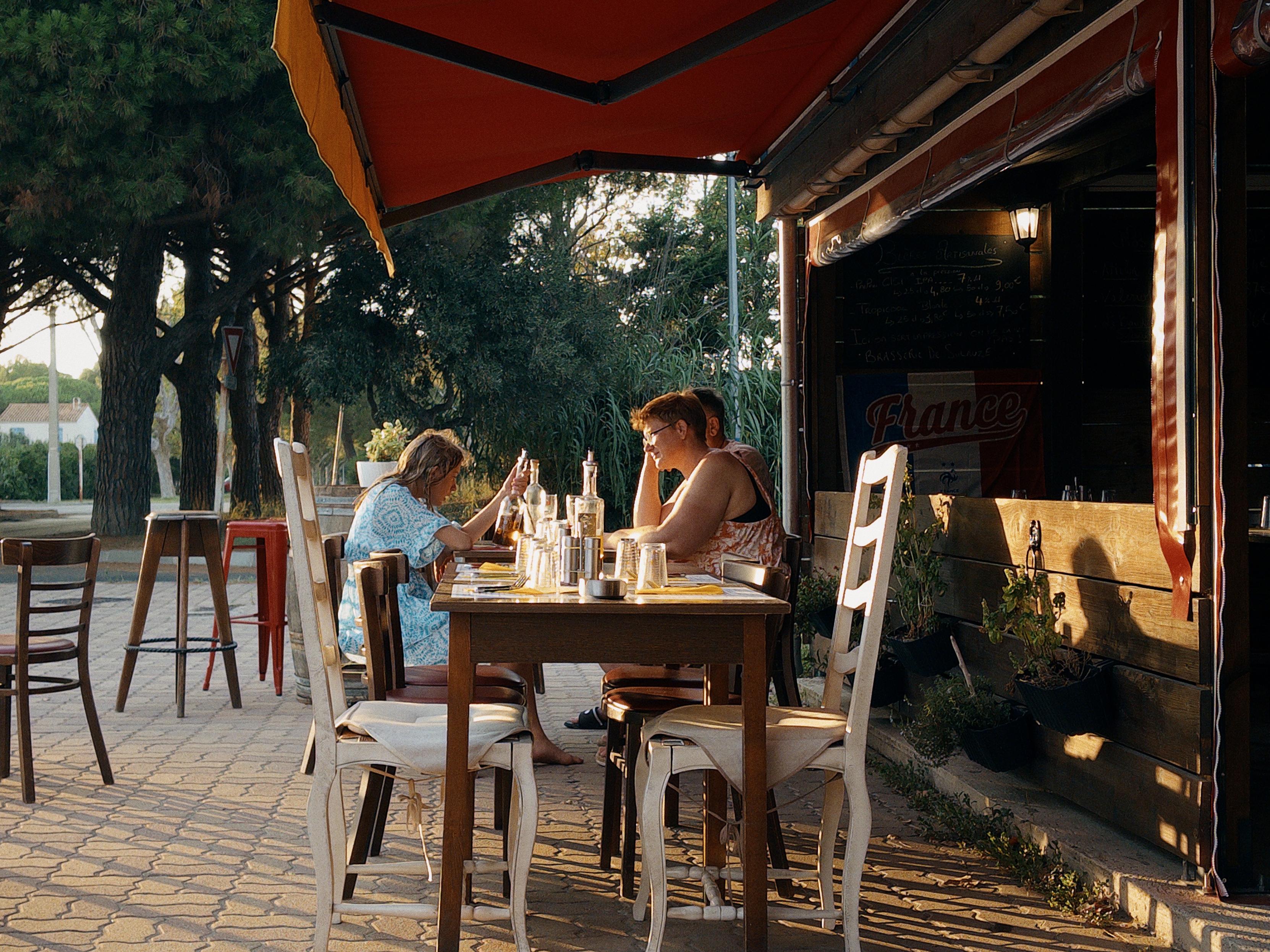 La foto mostra due persone sedute a un tavolo all'aperto in un bar o ristorante, illuminate dalla luce calda del tardo pomeriggio. Sono intenti a guardare i loro telefoni, con bottiglie e bicchieri sparsi sul tavolo. L'ambiente è accogliente, con tavoli e sedie in legno, un tendone rosso che copre parzialmente la terrazza, e alberi sullo sfondo che creano un'atmosfera rilassata e ombreggiata.