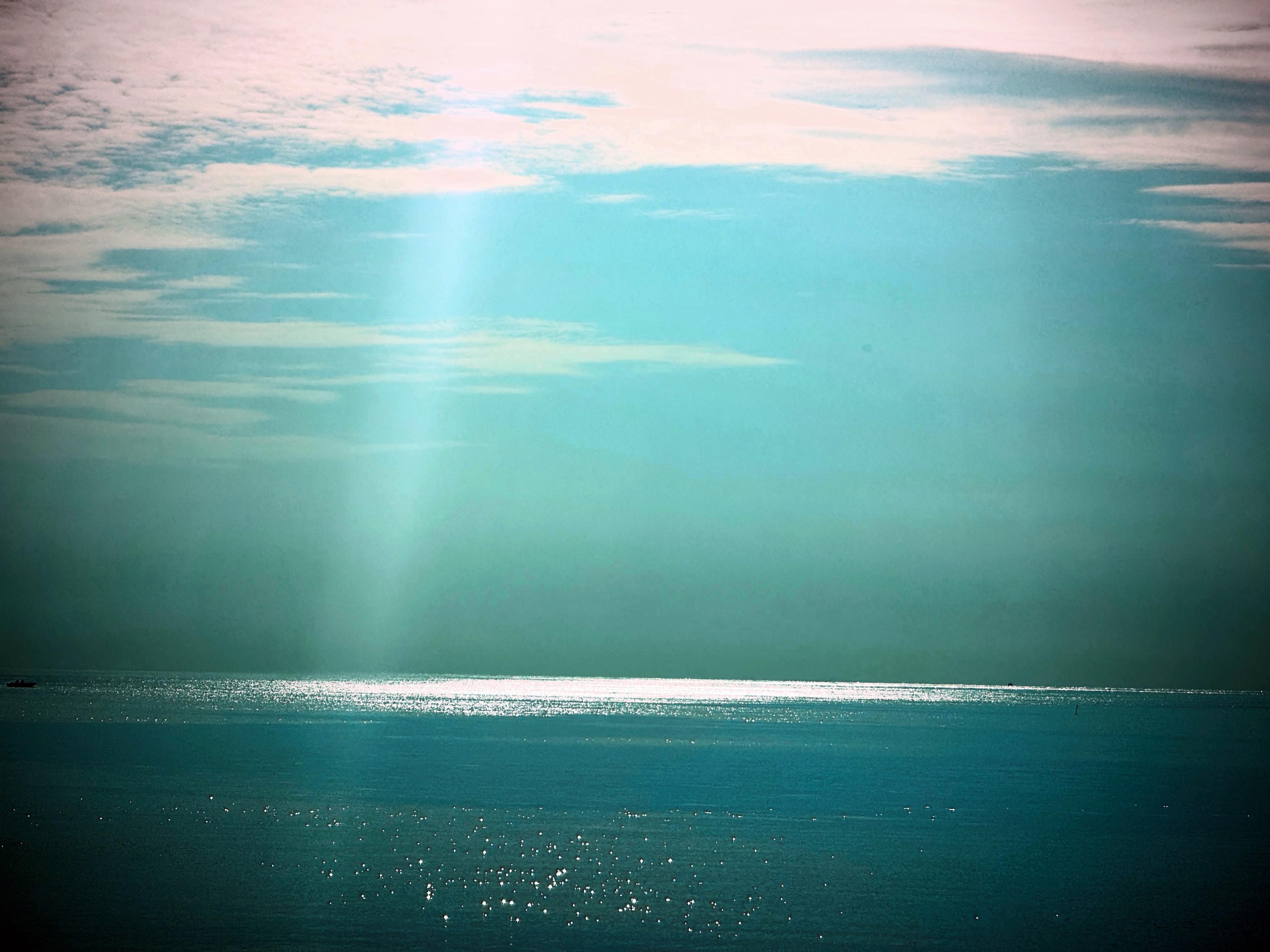 Mare calmo sotto un cielo con nuvole leggere e un fascio di luce solare che si riflette sull'acqua.